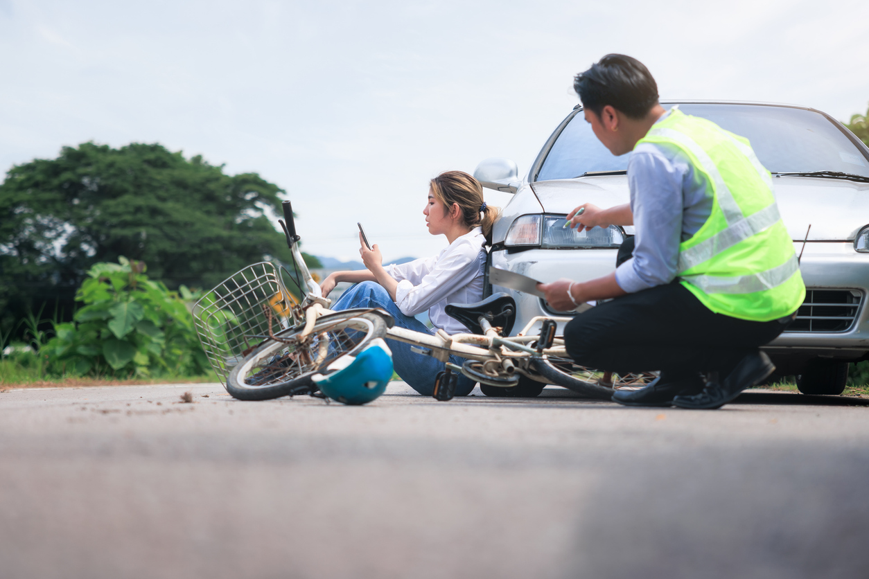 Injured bicyclist sitting on the roadside after a collision with a car while another person documents the accident details
