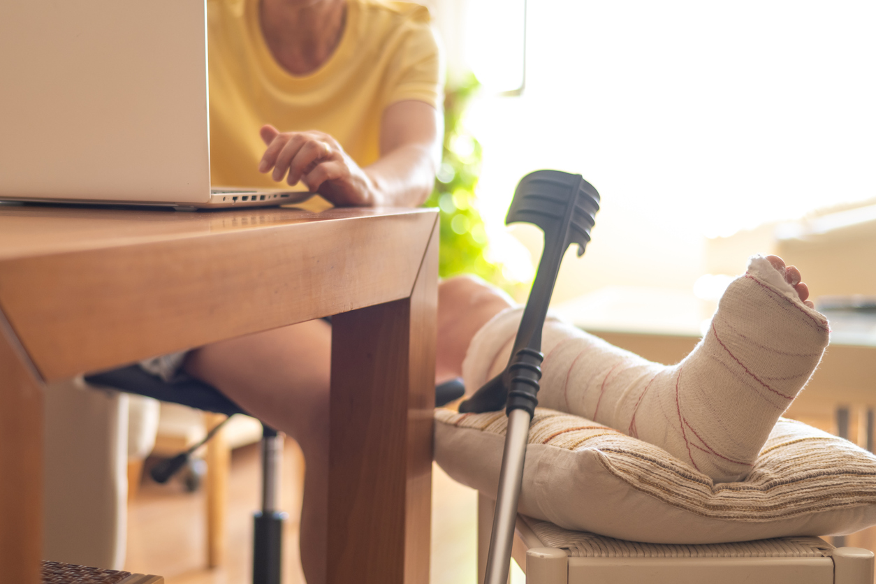 Person working on a laptop at home with a leg in a cast and crutches nearby after being injured outside of work