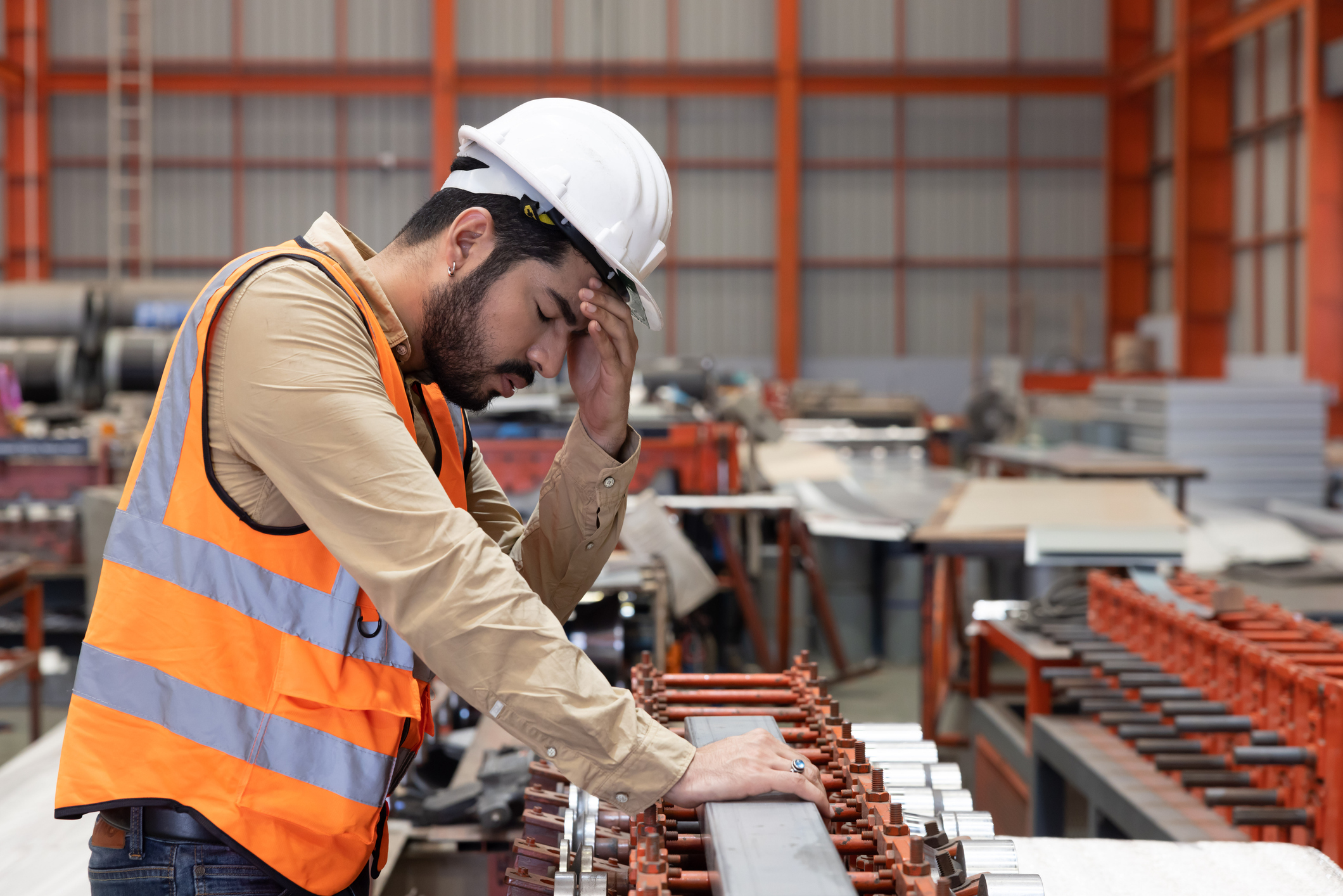 Factory worker wearing a hard hat and safety vest leaning over equipment with a hand on his head, representing stress or symptoms related to an occupational illness.