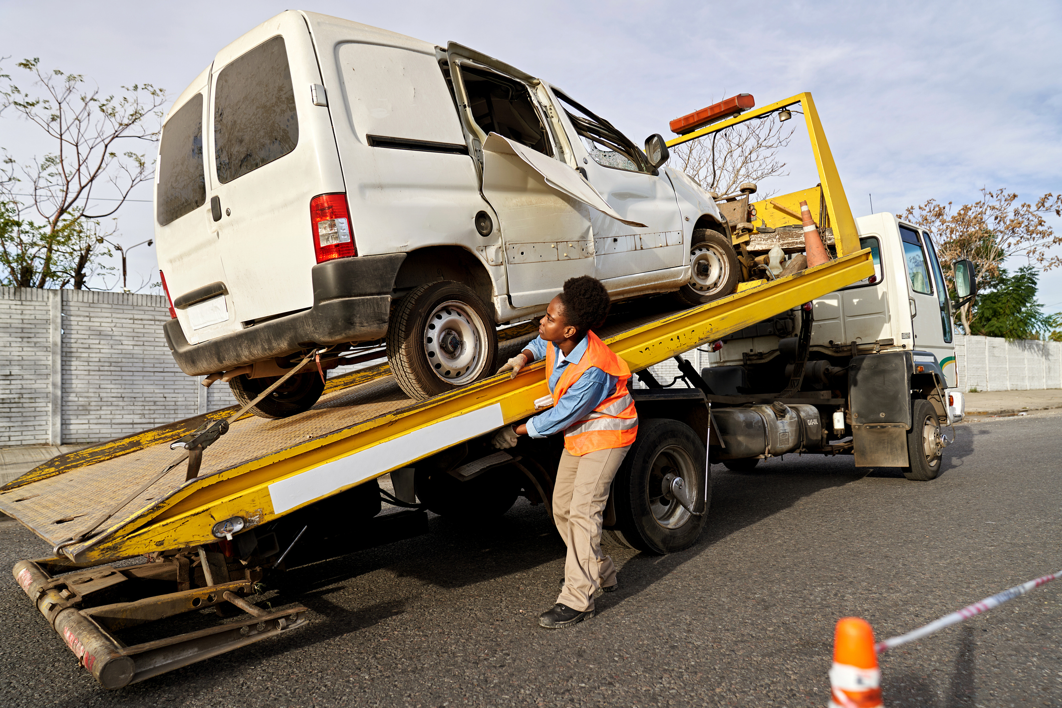 Operador de grúa cargando una camioneta muy dañada tras un accidente de vehículo relacionado con el trabajo, usando equipo de seguridad a un costado de la carretera.