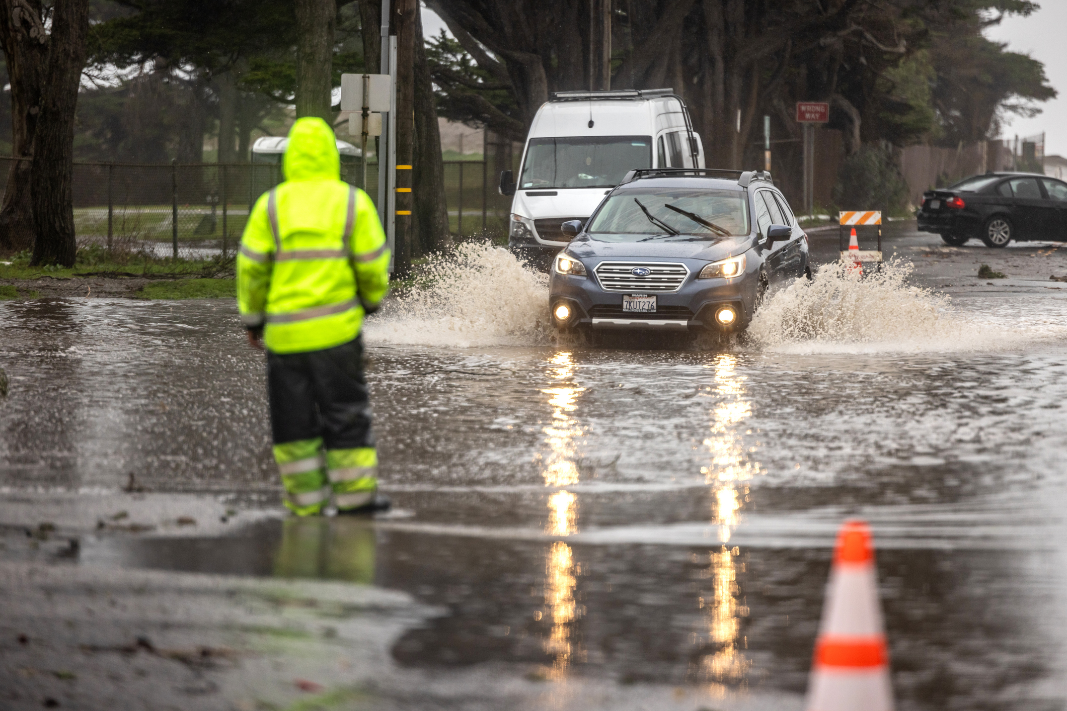 Car driving through a flooded roadway during heavy rain while a safety worker stands nearby, illustrating hazardous weather conditions
